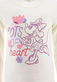 Light cream long-sleeve shirt featuring purple and pink graphic of a character with polka dots, text "LOTS OF heart," and playful flower accents.