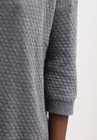 Pull en tricot gris avec un motif texturé, manches trois-quarts et poignets côtelés. Le tissu semble doux et de teneur moyenne.