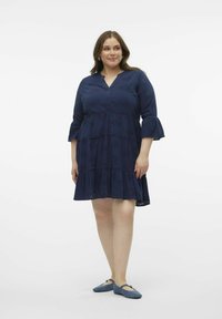 Dunkelblaues Baumwollkleid mit V-Ausschnitt, 3/4-Ärmeln im Glockenstil, schichtenweise Rüschen und Lochstickereien. Kombiniert mit blauen Ballerinas.