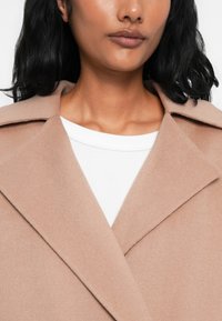 Cappotto di lana beige chiaro con un colletto e rever strutturati, caratterizzato da una texture morbida. Sotto, è visibile una maglietta bianca con colletto tondo.