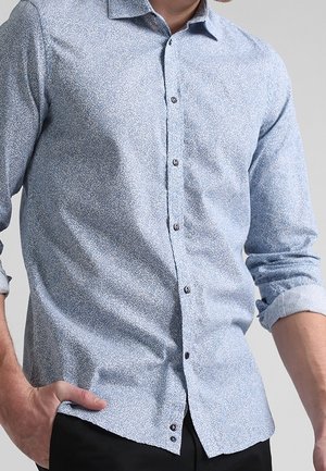 Mężczyzna w jasnoniebieskiej wzorzystej koszuli z długim rękawem, z podwiniętymi mankietami i czarnych spodniach, z ręką w kieszeni, na jednolitym tle.