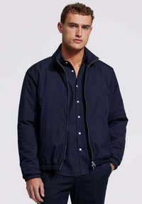 Marine blauwe sweatjacket met rits en elastische boorden en zoom, gemaakt van een gladde stof. Gepaard met een donker shirt met kraag, weergegeven op een neutrale achtergrond.