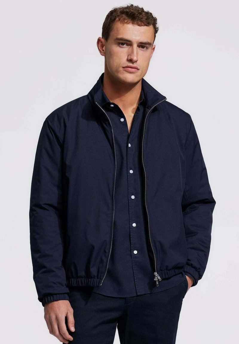 Marine blauwe sweatjacket met rits en elastische boorden en zoom, gemaakt van een gladde stof. Gepaard met een donker shirt met kraag, weergegeven op een neutrale achtergrond.