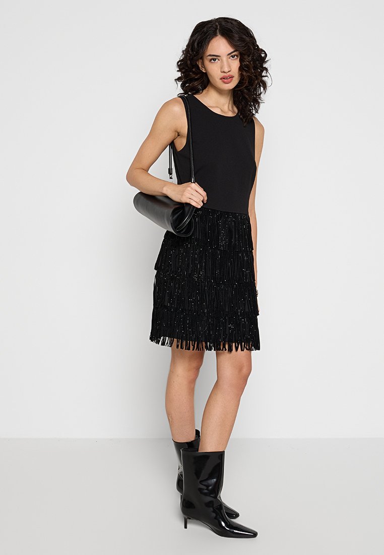 Czarna sukienka bez rękawów z frędzlami u dołu. Teksturowany materiał z cekinami, zestawiona z błyszczącymi czarnymi kozakami do kolan i okrągłą torebką.
