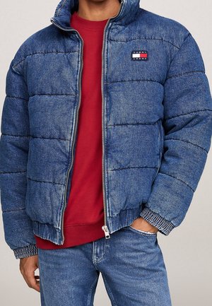 Homme portant une veste en jean matelassée bleu Tommy Jeans par-dessus un sweat-shirt rouge, une main dans la poche du jean.