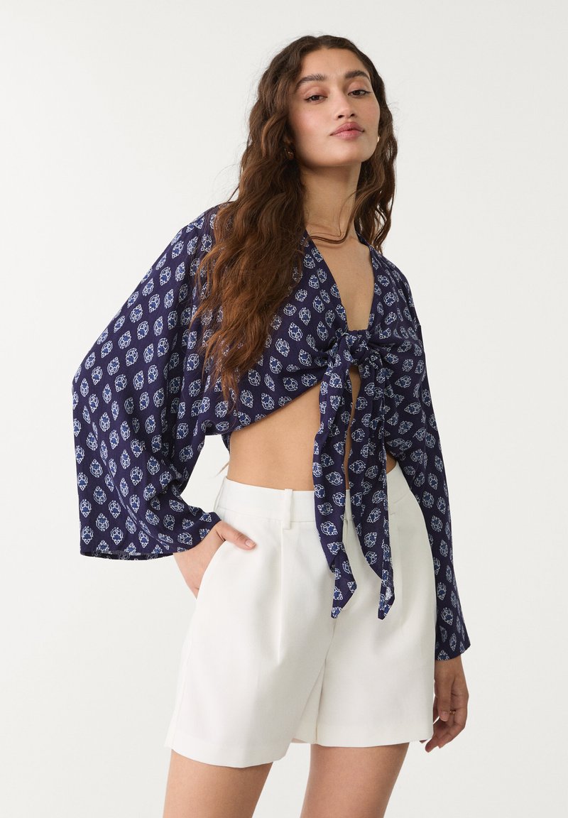Un haut court bleu marine avec un motif floral blanc complexe, noué à l'avant. Associé à un short blanc taille haute. Manches amples et fluides.