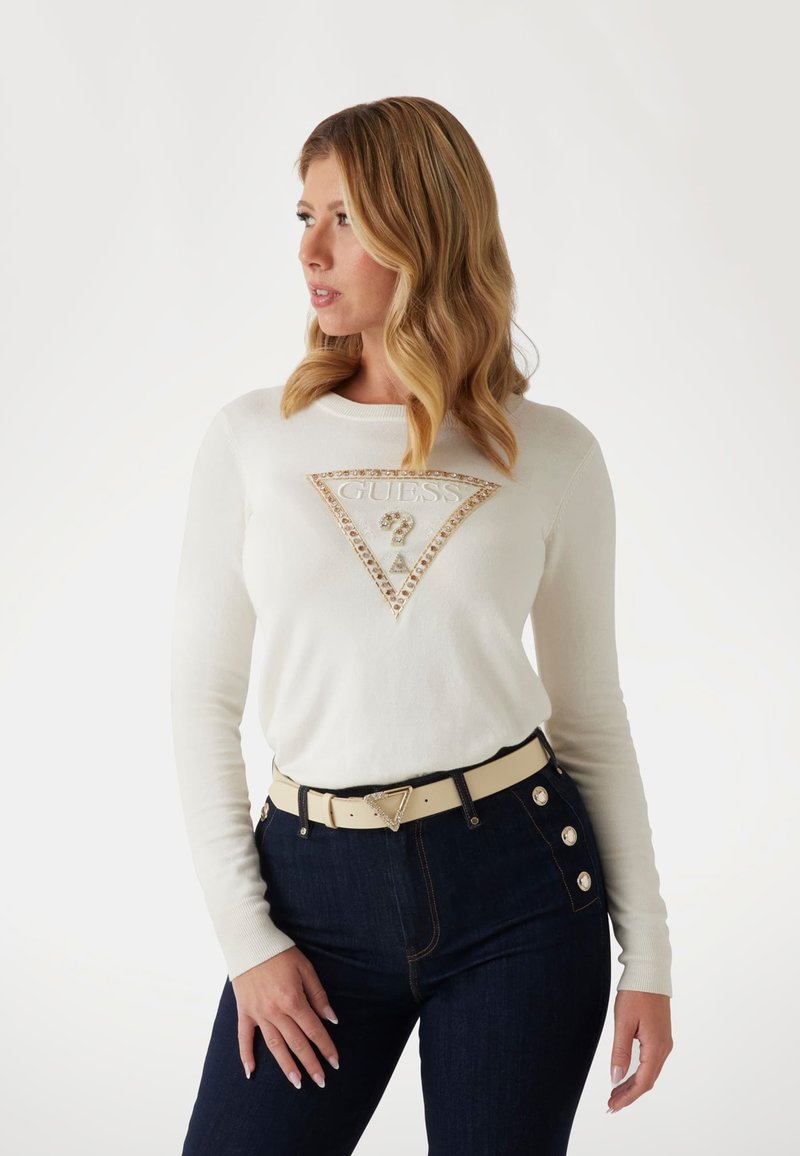 Suéter blanco de manga larga con un logo de strass en un diseño triangular. Combinado con jeans de mezclilla oscuros que tienen botones decorativos. Cinturón beige incluido.