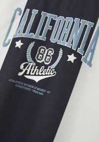 Sports T-skjorte med et svart og hvitt fargeblokkmønster, med ordet "CALIFORNIA" i fet lys blå skrift og grafiske detaljer.