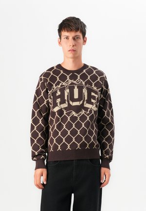 Jeune homme portant un pull marron avec un motif en maillons beiges et un texte "FUEL" en gras, associé à un pantalon noir ample, debout devant un fond blanc.