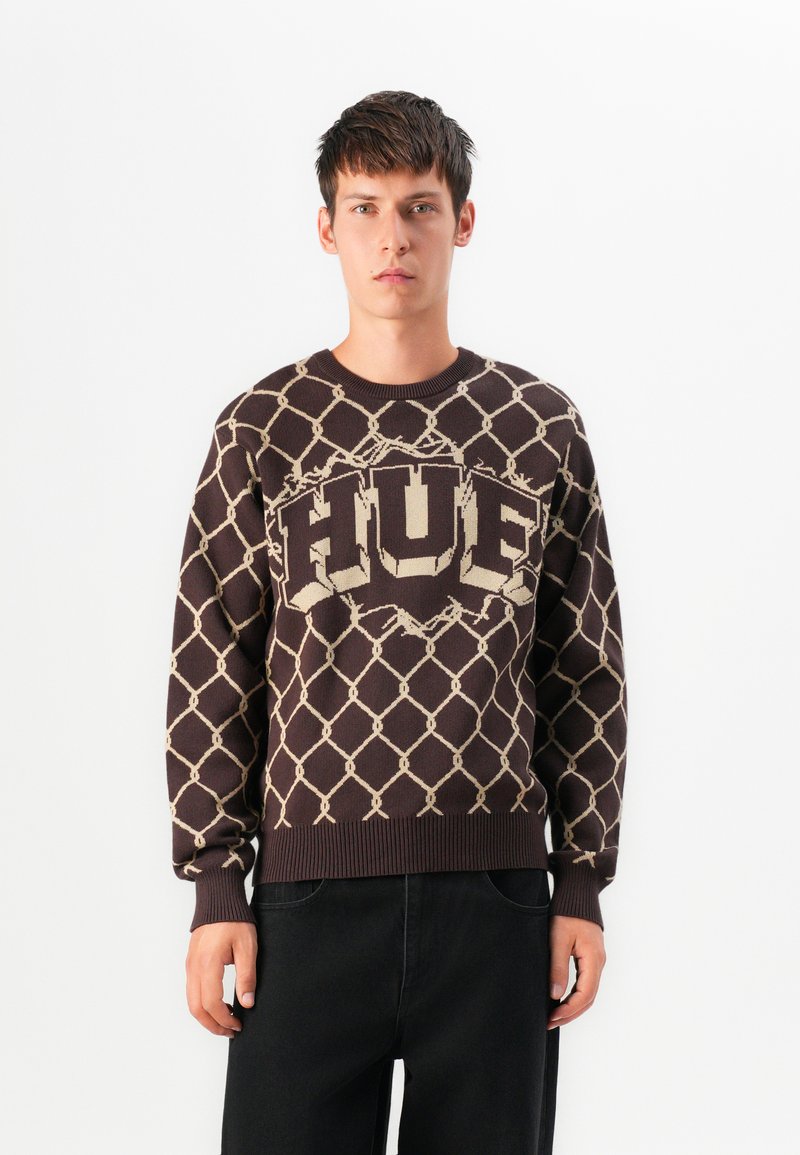 Young man wearing a brown sweater with beige chain-link pattern and bold "FUEL" text, paired with loose black pants, standing against white background.