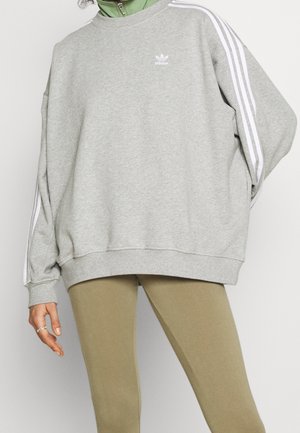 Osoba nosząca szarą bluzę Adidas z białymi paskami na rękawach i zielonym kołnierzem, w połączeniu z jasnobrązowymi legginsami.