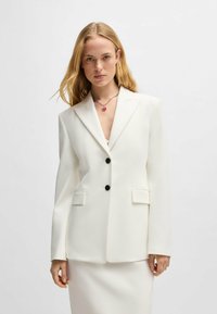 White tailored blazer featuring two buttons, peak lapels, side pockets, and a structured silhouette. Smooth texture with a clean finish.