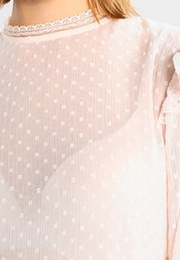 Przezroczysta różowa bluzka o kroju z kropkowaną fakturą, z koronkowym wykończeniem dekoltu i falbaniastymi rękawami, oferująca miękki, lekki wygląd.
