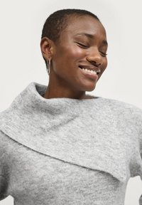 Grauer Pullover mit weitem Kragen, aus weichem, strukturiertem Strick und asymmetrischem Design. Das Model trägt Ohrstecker.