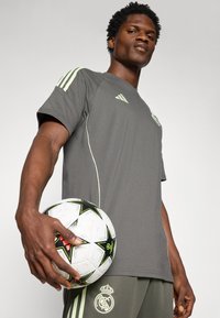 Grå sports T-shirt med lysegrønne detaljer og logo, parret med grå bukser med en klubemblem. Holder en sort og hvid fodbold.