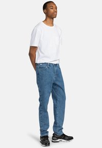 Witte katoenen T-shirt met een zak, gecombineerd met blauwe spijkerbroeken. Model draagt zwarte sneakers met textuurdetails en staat in profiel.