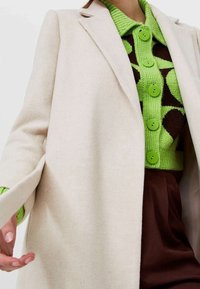 Manteau en laine beige sur un cardigan à motifs verts avec de grands boutons ; pantalons marron visibles ; texture lisse et design ajusté.