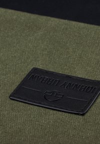 Olívazöld szövet folt fekete, texturált bőrcímkével, amelyen a "Johnny Urban" felirat domborítva, valamint egy kör alakú logó részlet található.