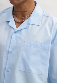 Camicia azzurra chiara con bottoni, realizzata in tessuto morbido, con colletto classico, taschino sul petto e bottoni azzurri chiari per la chiusura.