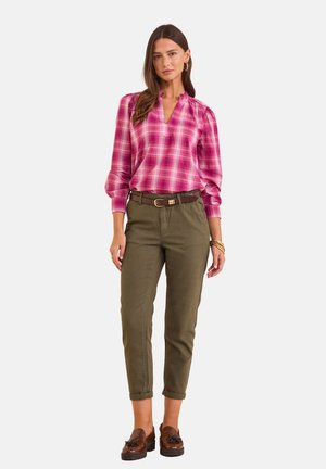 Femme portant un chemisier à manches longues à carreaux roses, un pantalon ceinturé vert olive, des mocassins marron et des bracelets dorés, debout sur un fond blanc.