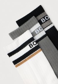 Trzy pary skarpet w kolorze białym, szarym i czarnym. Każda ma poziome paski z logo tekstowym. Materiał to miękka, prążkowana dzianina.