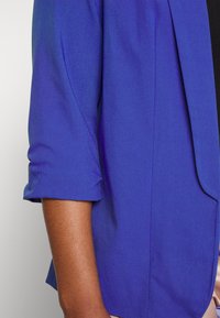 Brazo y torso vistiendo un blazer azul real con las mangas arremangadas, mostrando un tono de piel marrón medio.