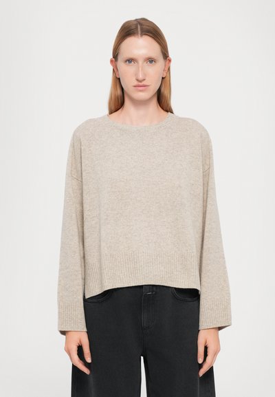 Femme avec des cheveux raides clairs portant un pull à manches longues beige à côtes et un pantalon noir ample, se tenant devant un fond blanc uni.