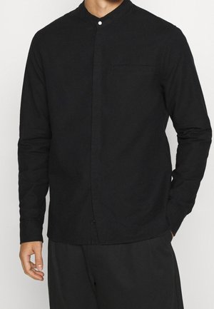 Homme portant une chemise noire à manches longues avec col mandarin et boutons cachés, ainsi qu'un pantalon noir assorti, main partiellement dans la poche.