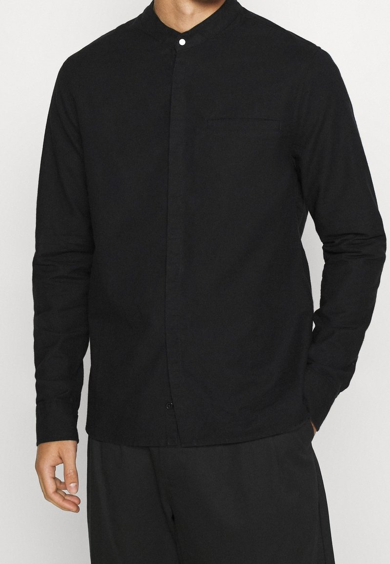 Homme portant une chemise noire à manches longues avec col mandarin et boutons cachés, ainsi qu'un pantalon noir assorti, main partiellement dans la poche.