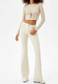 Beige Langarm-Crop-Top mit einer zentralen Krawatten-Detail und hell gefärbten, ausgestellten Jeans. Glatte Textur, figurbetontes Design. Schlichter Stil.