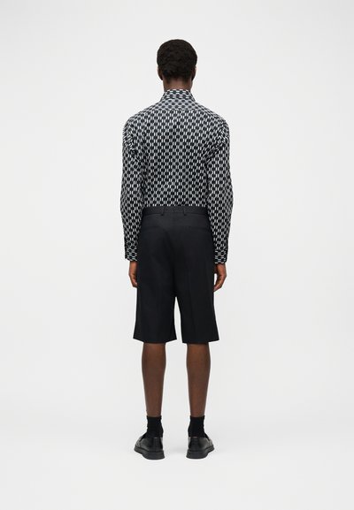 Homme debout, de dos, portant une chemise à manches longues noire et blanche à motifs, un short noir jusqu'aux genoux, des chaussettes noires et des chaussures noires.