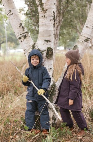 Deux enfants se tiennent contre un bouleau. Le garçon porte un manteau à capuche bleu marine et des gants jaunes, tandis que la fille porte un manteau violet foncé et un chapeau marron.