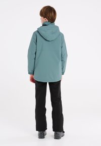 Veste imperméable teal avec capuche, poignets élastiques et ourlet droit, associée à un pantalon noir et des chaussures gris foncé.