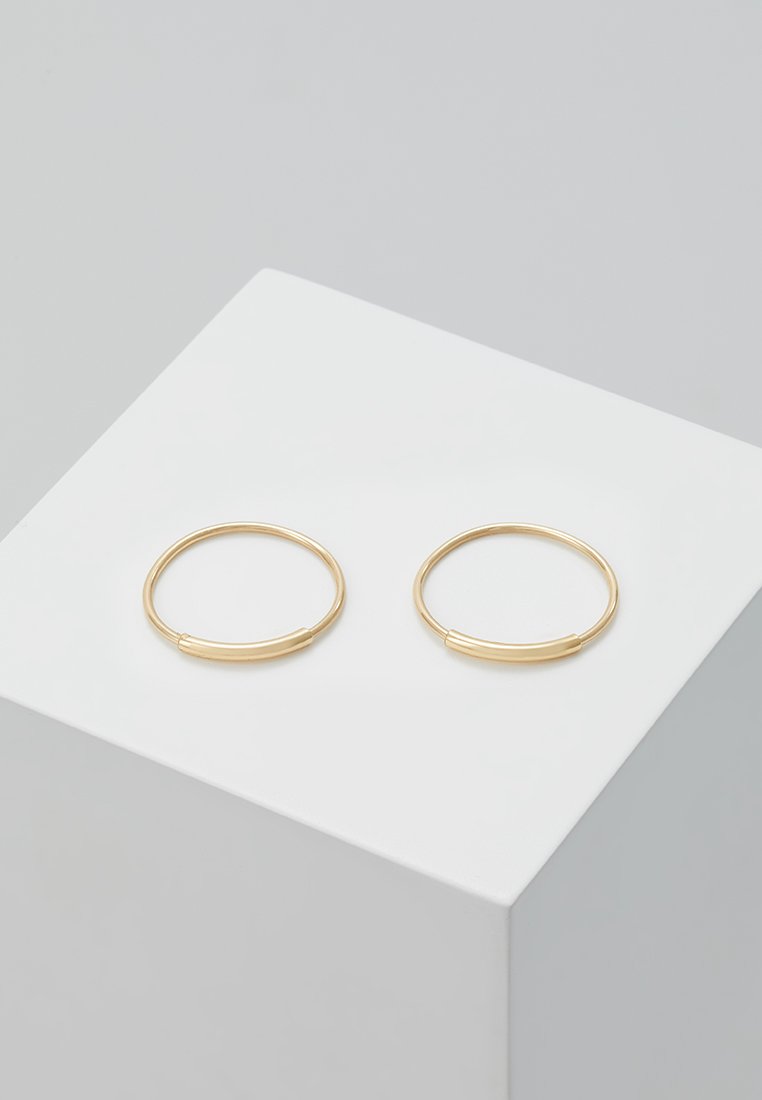 Pendientes de aro de oro con una forma redonda y lisa, con un sutil acento de diseño. Colocados sobre un fondo blanco de exhibición.