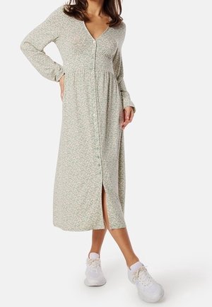 Robe à fleurs légère à manches longues, couleur vert clair, avec un décolleté en V, fermeture à boutons et une fente à l'avant, associée à des baskets blanches. Matière en mélange de coton.