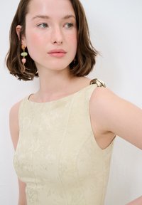 Beige mouwloze jurk met bloemtextuur, voorzien van een ronde metalen accent op de linkerschouder. Gladde stof met een aansluitende snit.