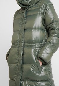 Persona vestida con una chaqueta de invierno acolchada de color verde oliva brillante, con las manos en los bolsillos delanteros, cierre de cremallera y botones a presión visibles.