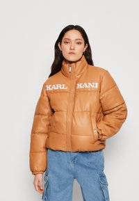 Piesková puffer bunda vyrobená z umelej kože, s vysokým golierom, bielym logom "KARL KANI" a elastickými manžetami. Predný zips a vrecká.