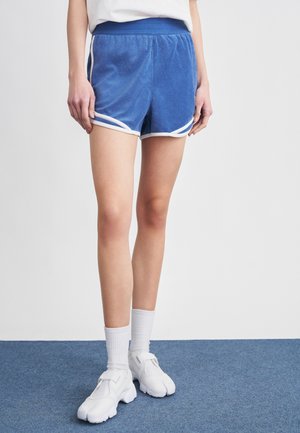 Egy személy kék frottír rövidnadrágot visel fehér szegéllyel, fehér középmagas zoknival és fehér, hasított orrú sportcipővel, aki kék szőnyegen áll.