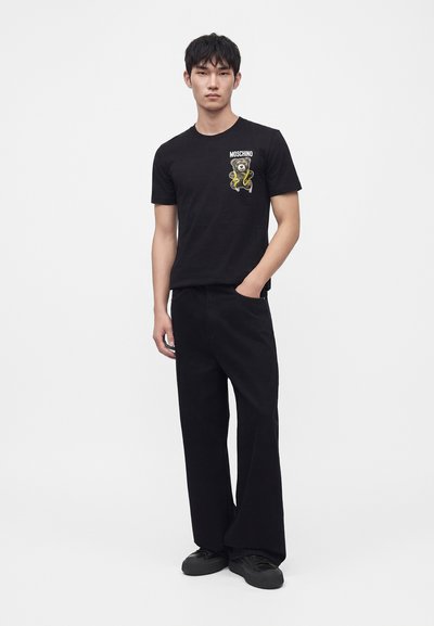 Νέος άνδρας στέκεται φορώντας μαύρο t-shirt Moschino με σχέδιο αρκουδάκι, μαύρο παντελόνι με ευρύ πατρόν και μαύρα αθλητικά, λευκό φόντο.
