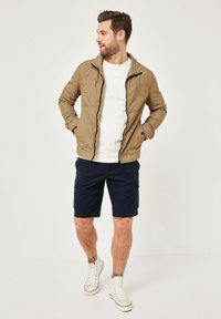 Tan lightweight jacket with a zip front, white T-shirt, and navy cargo shorts. Model wearing white sneakers with rolled cuffs.