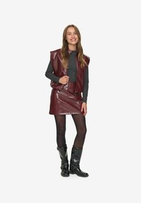 Donna sorridente, che indossa un gilet in pelle bordeaux e una gonna, un top a maniche lunghe grigio scuro, collant neri e stivali neri fino a metà polpaccio, in piedi su uno sfondo bianco.