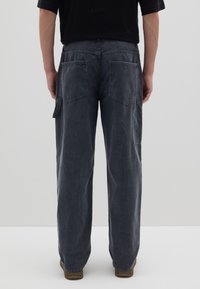 Graue Cordhosen mit geradem Schnitt, ausgestattet mit zwei Gesäßtaschen und einer Seitentasche. Braune Schuhe ergänzen das Outfit.