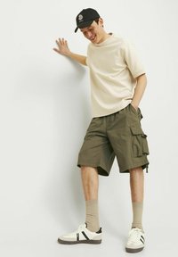 Beżowa koszula z krótkim rękawem, oliwkowe szorty cargo z kieszeniami, beżowe skarpetki i białe buty z czarnymi akcentami. Czarna czapka z detalem logo.