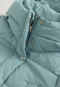 Gesteppte blaue Steppjacke mit kariertem Muster, ausgestattet mit einem Metallreißverschluss und Druckknöpfen vorne. Weiche Textur und gepolstertes Design.