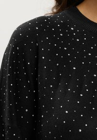 Suéter de punto negro con pequeños y medianos rhinestones plateados dispersos cerca del hombro y la parte superior del pecho.