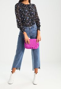 Donna con una blusa floreale nera, jeans Blu a vita corta con spacchi laterali, stivaletti bianchi alla caviglia, che tiene in mano una borsa a tracolla di un vivace colore rosa.