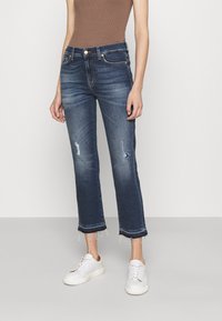 Jeans in denim blu scuro con gamba dritta, dettagli strappati, orlo sfrangiato e design a cinque tasche, abbinati a sneakers bianche.