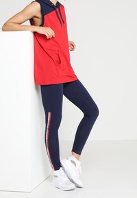 Person, der læner sig op ad en hvid blok, iført en ærmeløs rød og marineblå hoodie, marineblå leggings med sidelænstreg, og hvide sportssko.