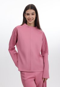 Rosa genser med lange ermer og blonderdetaljer på skuldrene, glatt tekstur og høy hals, sammen med koordinerte tettsittende bukser.